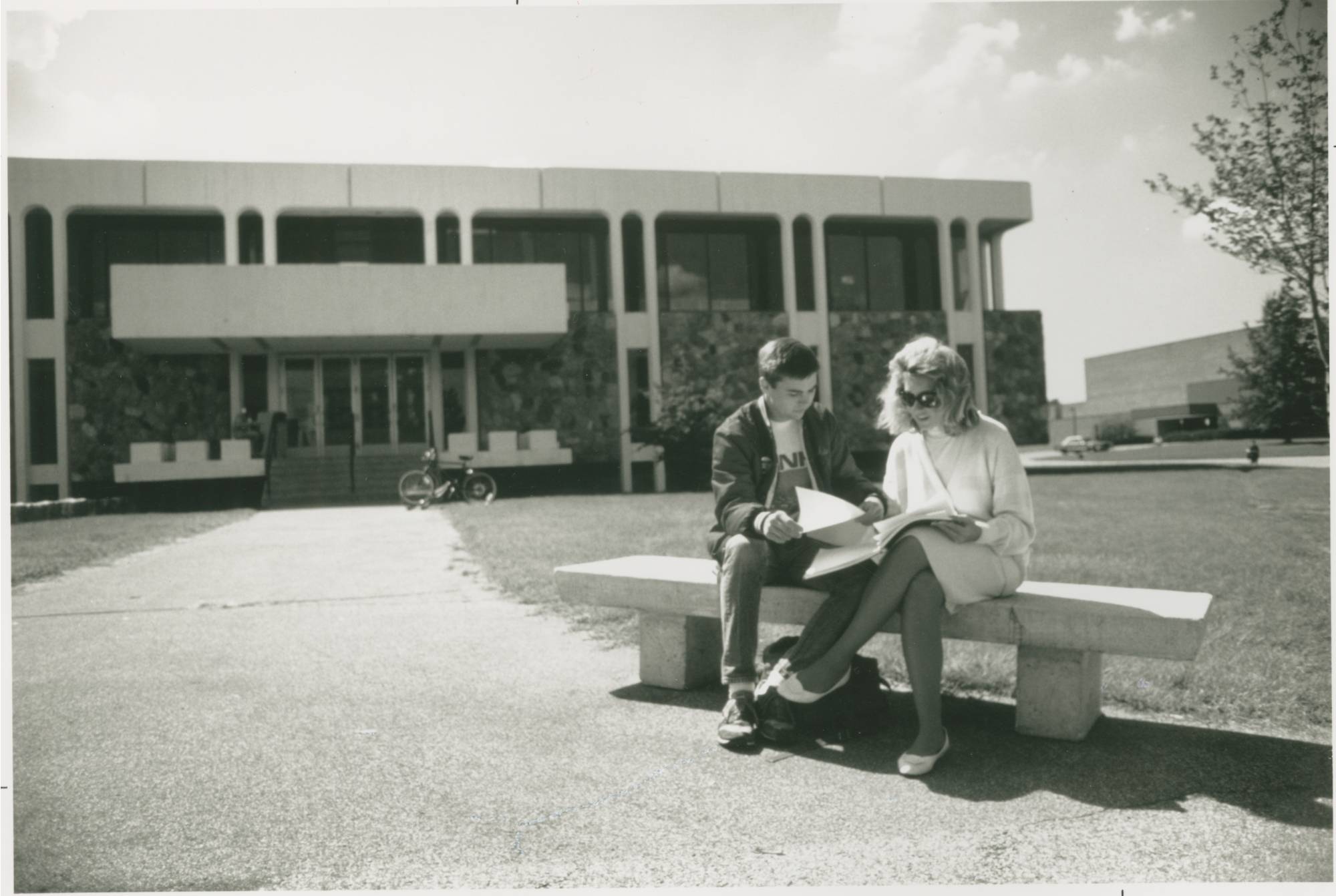 A black and white photo of a couple sitting on a bench in front of Lake Superior Hall on a sunny day.
